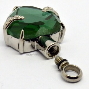 May include: A silver heart-shaped cremation pendant with a green crystal top. The pendant is open and the screw-on lid is separate.