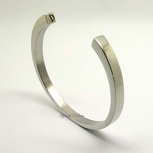 May include: A silver bangle bracelet with a flat, wide band. The bracelet has a screw closure.