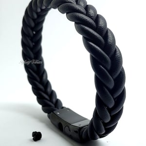 Könnte beinhalten: Schwarzes geflochtenes Lederarmband mit einem schwarzen Metallverschluss. Das Armband liegt auf einem weißen Hintergrund.