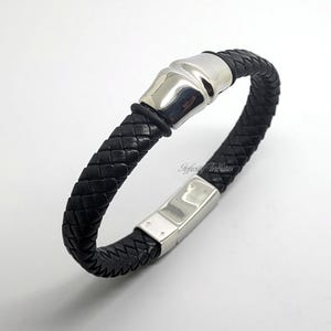Könnte beinhalten: Schwarzes geflochtenes Lederarmband mit silberfarbenem Verschluss und Akzenten. Das Armband hat ein gewebtes Design und einen Magnetverschluss. Ein stilvolles Accessoire.