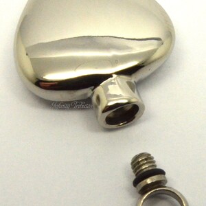 May include: A silver heart-shaped cremation urn with a threaded screw-on lid and a separate silver ring for attaching to a chain. The urn is designed to hold a small amount of ashes or other mementos.