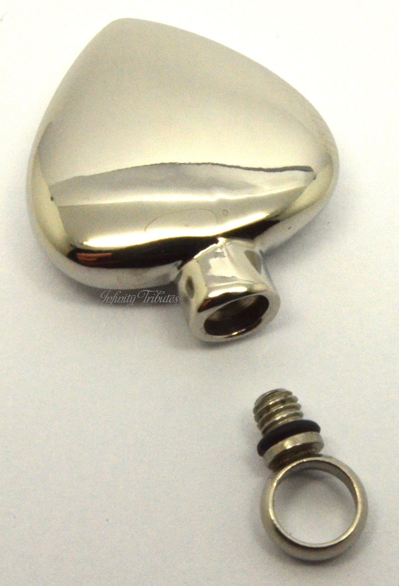 May include: A silver heart-shaped cremation urn with a threaded screw-on lid and a separate silver ring for attaching to a chain. The urn is designed to hold a small amount of ashes or other mementos.