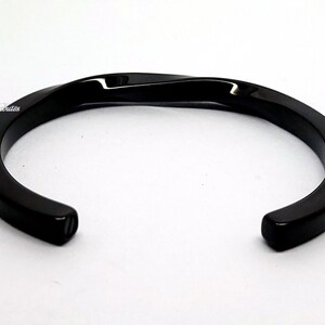 Könnte beinhalten: Schwarzes, gedrehtes Metall-Armband. Das Armband ist offen und hat eine glatte, polierte Oberfläche.