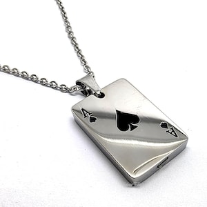 Puede incluir: Collar con colgante plateado que presenta el diseño de la carta del As de Picas. El colgante rectangular tiene un acabado pulido y está suspendido de una delicada cadena.