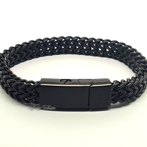Può includere: Un bracciale a catena di grano nero con una chiusura magnetica rettangolare liscia. Le maglie metalliche sono intrecciate in un motivo coerente e ripetitivo. Il testo Infinity Tributes è stampato in un carattere piccolo e chiaro vicino alla chiusura.
