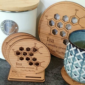 May include: Set of six wooden coasters with a chemical structure of tea printed on them. The coasters are round and have a light brown color. The text on the coasters reads "Tea, Camellia sinensis, C50 H50 N4 O26".