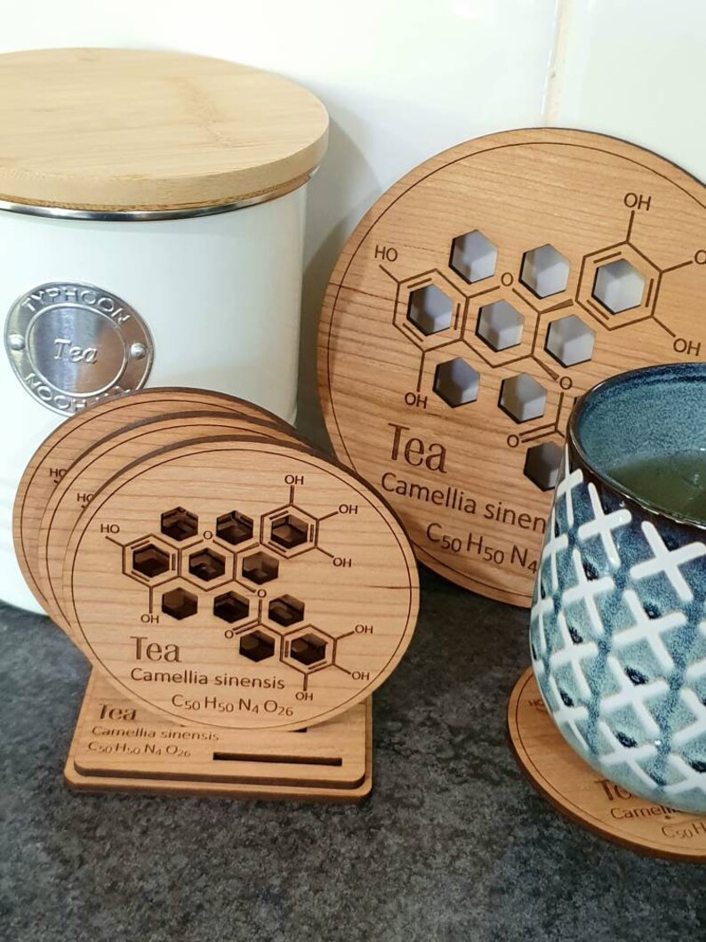 May include: Set of six wooden coasters with a chemical structure of tea printed on them. The coasters are round and have a light brown color. The text on the coasters reads "Tea, Camellia sinensis, C50 H50 N4 O26".