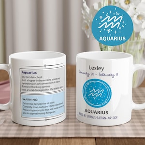 May include: Two white ceramic mugs featuring Aquarius zodiac designs. One mug displays a definition of Aquarius and a warning label. The other mug shows the name Lesley, the date range January 20-February 18, and a blue circular Aquarius icon.