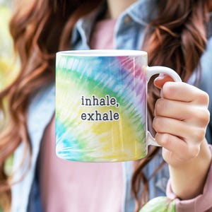 May include: A white ceramic mug featuring a vibrant, multi-colored tie-dye pattern in shades of blue, green, yellow, and pink. The text "inhale, exhale" is printed in a simple, black, sans-serif font on the center of the mug.