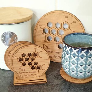 May include: Set of four wooden coasters with a chemical formula for tea printed on them. The coasters are round and have a light brown color. The formula is surrounded by a ring of text that reads "Tea Camellia sinensis C50 H50 N4 O26".