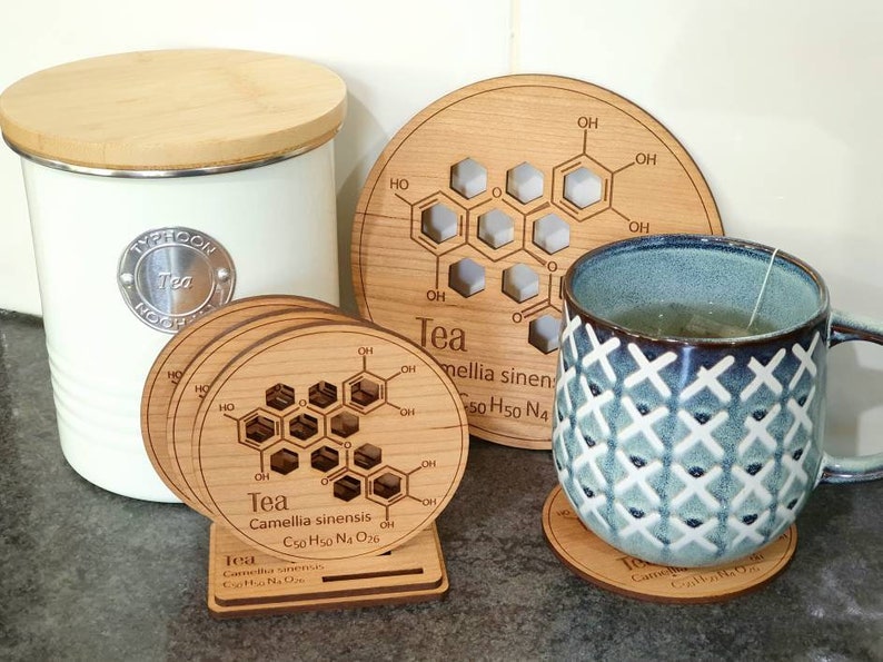 May include: Set of four wooden coasters with a chemical formula for tea printed on them. The coasters are round and have a light brown color. The formula is surrounded by a ring of text that reads "Tea Camellia sinensis C50 H50 N4 O26".