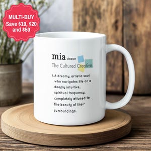 May include: A white ceramic mug featuring the printed definition of the name Mia, described as a cultured creative. The text includes a noun classification and a descriptive paragraph about an artistic soul, accompanied by three small colourful square icons.
