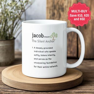 May include: A white ceramic mug featuring black text that defines the name Jacob as a noun, describing a deeply grounded individual who speaks softly, listens intently, and serves as the unwavering foundation for their entire network, with a tree icon.