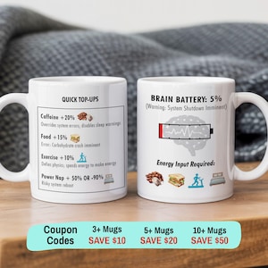 May include: Two white ceramic mugs featuring black text and colourful icons. One mug displays a list titled QUICK TOP-UPS with caffeine, food, exercise, and power nap options. The other mug shows a low battery icon over a brain with the text BRAIN BATTERY: 5%.