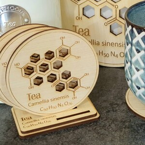 May include: Set of four wooden coasters with a chemical structure of tea printed on them. The coasters are stacked and have a stand. The text on the coasters reads "Tea Camellia sinensis C50 H50 N4 O26".