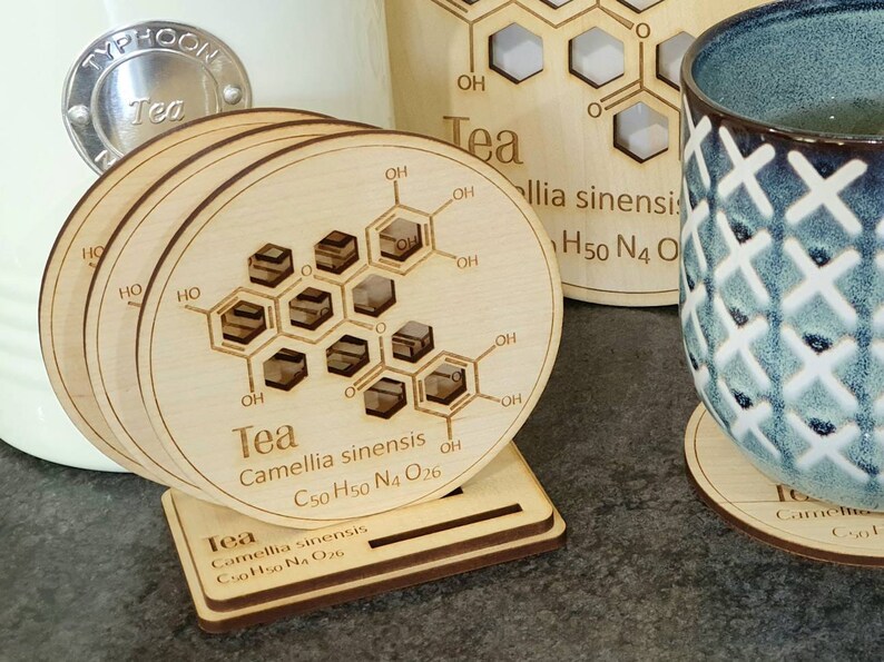 May include: Set of four wooden coasters with a chemical structure of tea printed on them. The coasters are stacked and have a stand. The text on the coasters reads "Tea Camellia sinensis C50 H50 N4 O26".