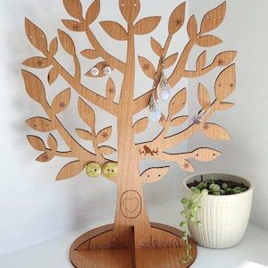 May include: Wooden tree-shaped earring holder with a heart carved into the trunk. The tree has multiple branches with holes for earrings. There are several pairs of earrings on the tree, including a pair of yellow and green button earrings, a pair of clear crystal earrings, and a pair of long, iridescent earrings. The tree is standing on a round base with a carved design.