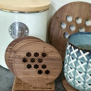 May include: Four wooden coasters with a chemical structure of tea printed on them. The coasters are stacked on a wooden stand. The chemical formula for tea is C50 H50 N4 O26. The coasters are made of walnut wood.