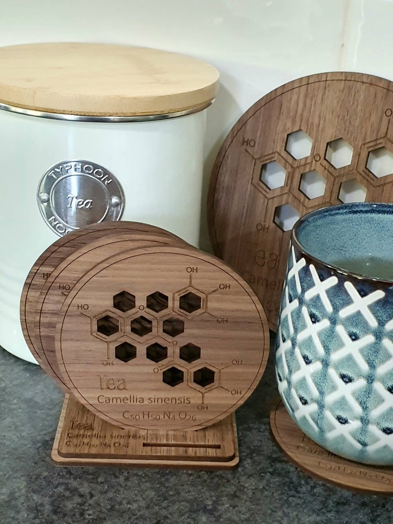 May include: Four wooden coasters with a chemical structure of tea printed on them. The coasters are stacked on a wooden stand. The chemical formula for tea is C50 H50 N4 O26. The coasters are made of walnut wood.