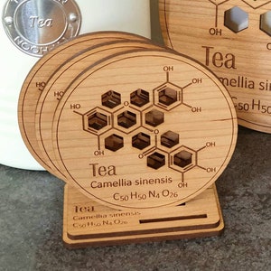 May include: Set of four wooden coasters with a chemical structure of tea printed on them. The coasters are stacked and have a small wooden stand. The text on the coasters reads "Tea Camellia sinensis C50 H50 N4 O26".