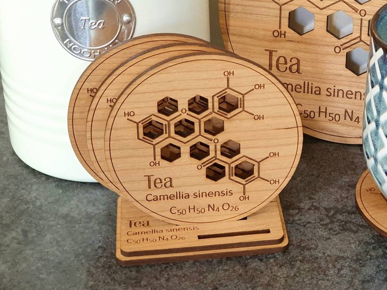 May include: Set of four wooden coasters with a chemical structure of tea printed on them. The coasters are stacked and have a small wooden stand. The text on the coasters reads "Tea Camellia sinensis C50 H50 N4 O26".