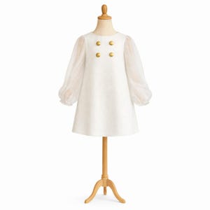 May include: A white dress with long, sheer sleeves and four gold buttons on the bodice. The dress is displayed on a wooden mannequin, showcasing its A-line silhouette. The dress is a classic design, suitable for special occasions.