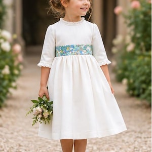 Peut inclure: Une jeune fille porte une robe blanche avec un bandeau floral et une ceinture bleue à motifs. Elle tient un petit bouquet de fleurs et porte des chaussures Mary Jane blanches. La robe a des manches courtes et un col à volants.