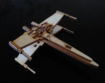 May include: A wooden model of an X-Wing starfighter from the Star Wars franchise. The model is made of light brown wood and has a detailed design.