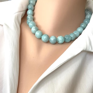 Op de afbeelding: Een lichtblauwe kralenketting met grote, ronde kralen. De ketting wordt gedragen op een witte top met kraag. De kralen hebben een licht gevlekt uiterlijk en de ketting lijkt een choker-stijl te zijn.