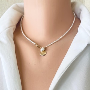 May include: A delicate necklace featuring a strand of small white seed pearls. The necklace includes a gold-toned metal seashell charm paired with a single white freshwater pearl accent, positioned as a pendant at the centre of the pearl strand.