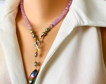 Pode incluir: Um colar longo e em camadas com um grande pingente de pérola iridescente. O colar é feito com contas de lavanda e verde e detalhes dourados.