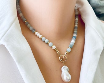 Puede incluir: Un collar delicado con un degradado de cuentas azules y grises, acentuado con perlas blancas y un gran colgante de perla irregular. El collar presenta un cierre dorado y una cadena dorada.