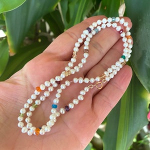 May include: A white pearl necklace with colourful gemstone accents. The necklace is made of small, round pearls and features a variety of gemstones in different colours, including red, orange, yellow, green, blue, and purple.