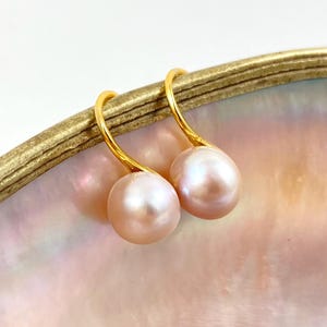 May include: A pair of drop earrings featuring two round, light pink pearls suspended from curved, polished gold-tone metal hooks. The pearls exhibit a smooth, lustrous surface and are positioned side by side, showcasing their soft, iridescent hue.