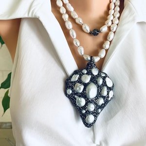 May include: A two-strand pearl necklace with a large, ornate pendant. The pendant is black with white pearl accents. The necklace is worn over a white collared shirt.