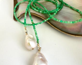 Pode incluir: Um colar de contas verdes com duas grandes pérolas barrocas brancas em forma de pêra. O colar é colocado sobre uma concha iridescente com borda dourada. As pequenas contas criam um design delicado e elegante.