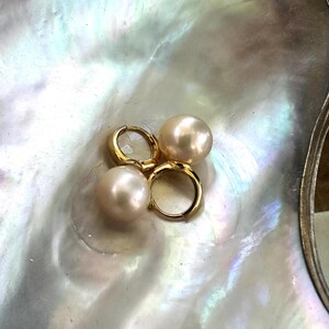 May include: A pair of gold hoop earrings, each featuring a single large, round, pale pinkish-white pearl attached to the bottom. The pearls have a smooth, lustrous surface, and the hoops are made of polished gold metal with a secure hinged closure.