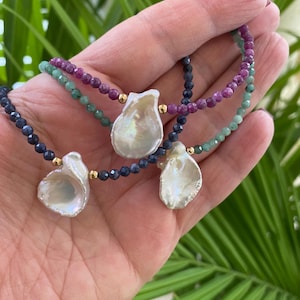 May include: Three necklaces with pearl pendants and gemstone beads. The necklaces feature a large, irregular-shaped white pearl pendant. The beads are in shades of blue, green, and purple, with gold accents.