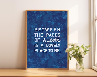 Könnte beinhalten: Ein gerahmter Kunstdruck mit blauem floralem Hintergrund und dem Zitat: "BETWEEN THE PAGES OF A book IS A LOVELY PLACE TO BE." Eine Vase mit weißen Blumen steht auf einer Holzoberfläche.