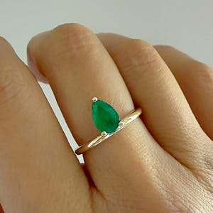 Op de afbeelding: Een gouden ring met een peervormige, levendige groene edelsteen. De ring heeft een slanke band en een kleine, ronde accentsteen. De ring wordt op een hand getoond.