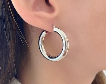 May include: Silver hoop earrings with a thick, rounded band. The earrings are worn in a person's ear.