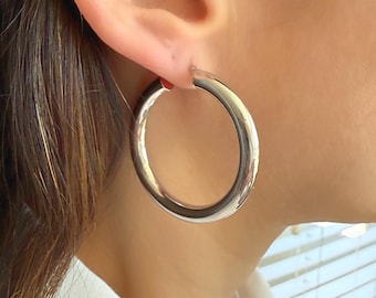 May include: A close-up view of a silver hoop earring. The earring is a thick, rounded design, reflecting light. The earring is worn on the ear of a person wearing a white collared shirt. The background is blurred.