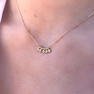 May include: A delicate gold necklace featuring a series of small, round beads and five clear, round gemstones. The necklace is displayed against a light background, highlighting the jewelry's fine details and elegant design.