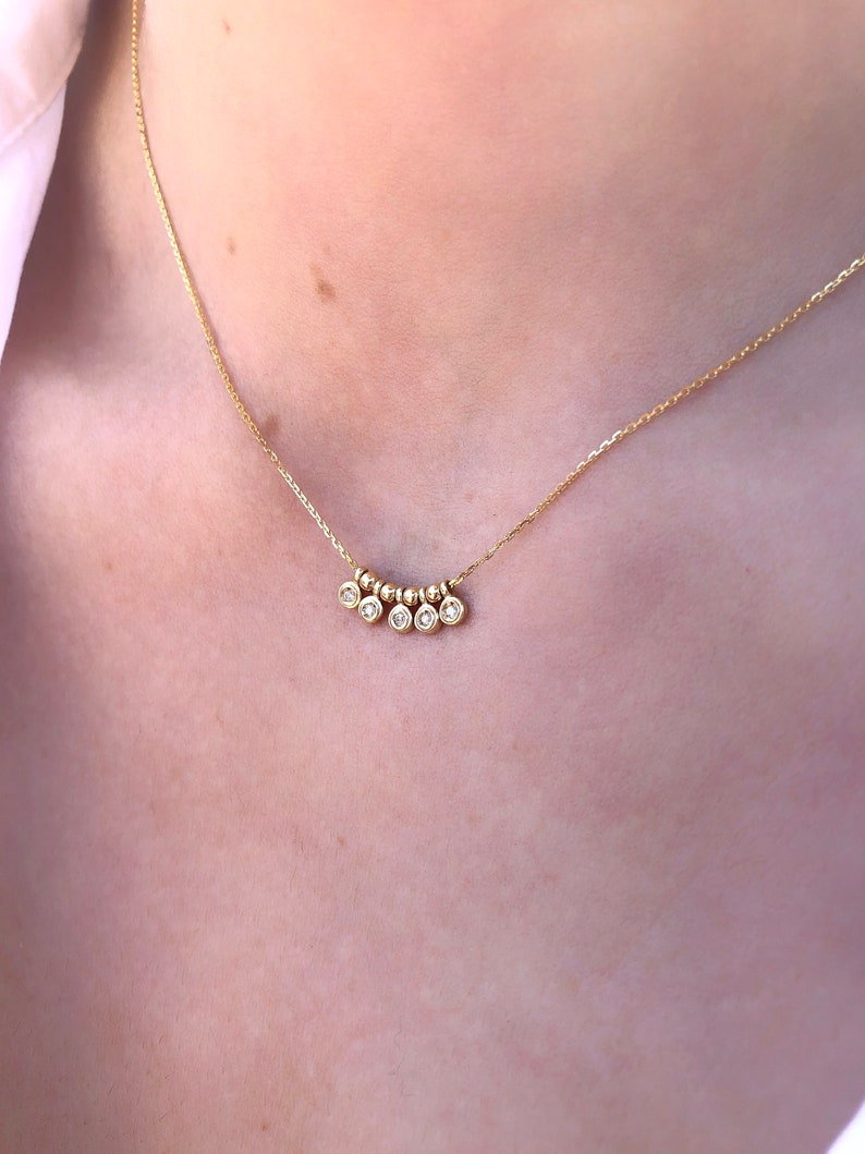 May include: A delicate gold necklace featuring a series of small, round beads and five clear, round gemstones. The necklace is displayed against a light background, highlighting the jewelry's fine details and elegant design.