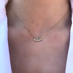 May include: A delicate gold necklace featuring five small, round, clear gemstones. The gemstones are set in a row, suspended from a thin gold chain. The necklace is worn against a white shirt.