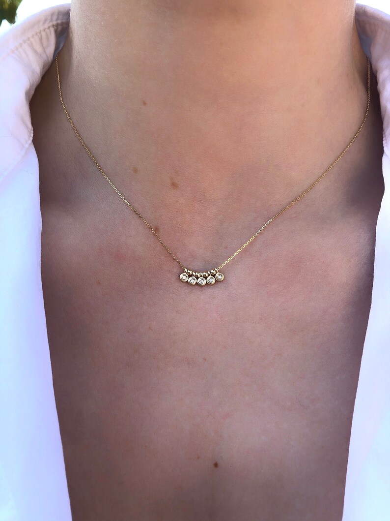 May include: A delicate gold necklace featuring five small, round, clear gemstones. The gemstones are set in a row, suspended from a thin gold chain. The necklace is worn against a white shirt.