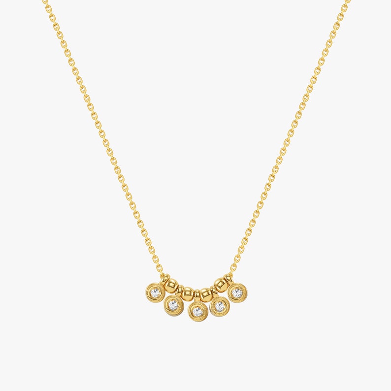 May include: A delicate gold necklace featuring a fine chain and a series of small, round beads. The necklace has a curved design with six bezel-set, clear gemstones. The overall design is elegant and minimalist.