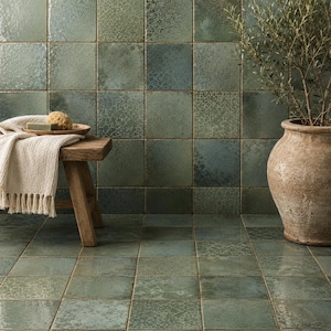 May include: Square ceramic tiles in shades of sage and forest green with a glossy, mottled finish cover the wall and floor. A small wooden stool holds a beige fringed cloth and a sponge, while a large terracotta pot sits on the floor.