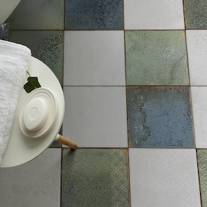 May include: A white round side table holds a stack of two white folded towels and a small white soap dish. The table sits on a floor covered in square ceramic tiles featuring a mix of solid white, mottled blue, and patterned sage green finishes.