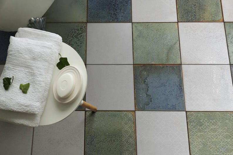 May include: A white round side table holds a stack of two white folded towels and a small white soap dish. The table sits on a floor covered in square ceramic tiles featuring a mix of solid white, mottled blue, and patterned sage green finishes.